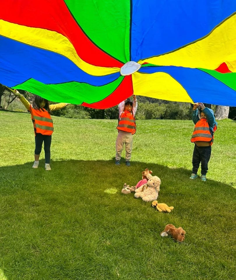 Children holding up a colorful parachute over stuffed animals on grass