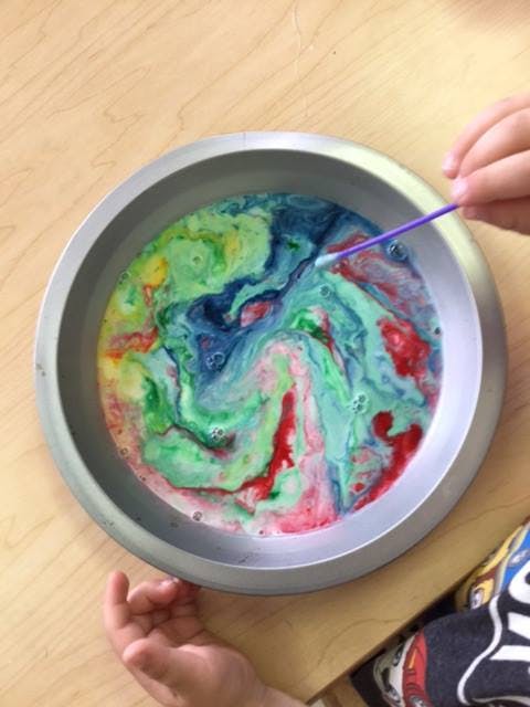 Child's hand using a cotton swab to mix colorful liquids in a pan