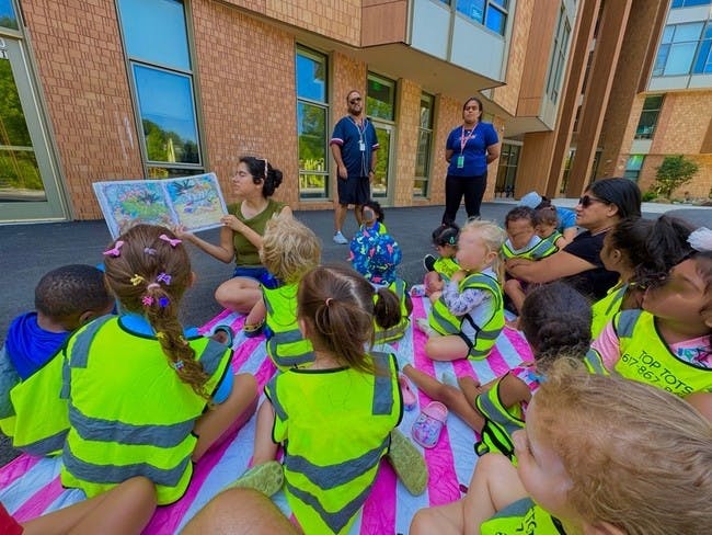 Adult reading a book to a group of children sitting on a blanket outdoors