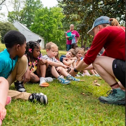 Adult person crouching and talking to a group of children sitting on grass