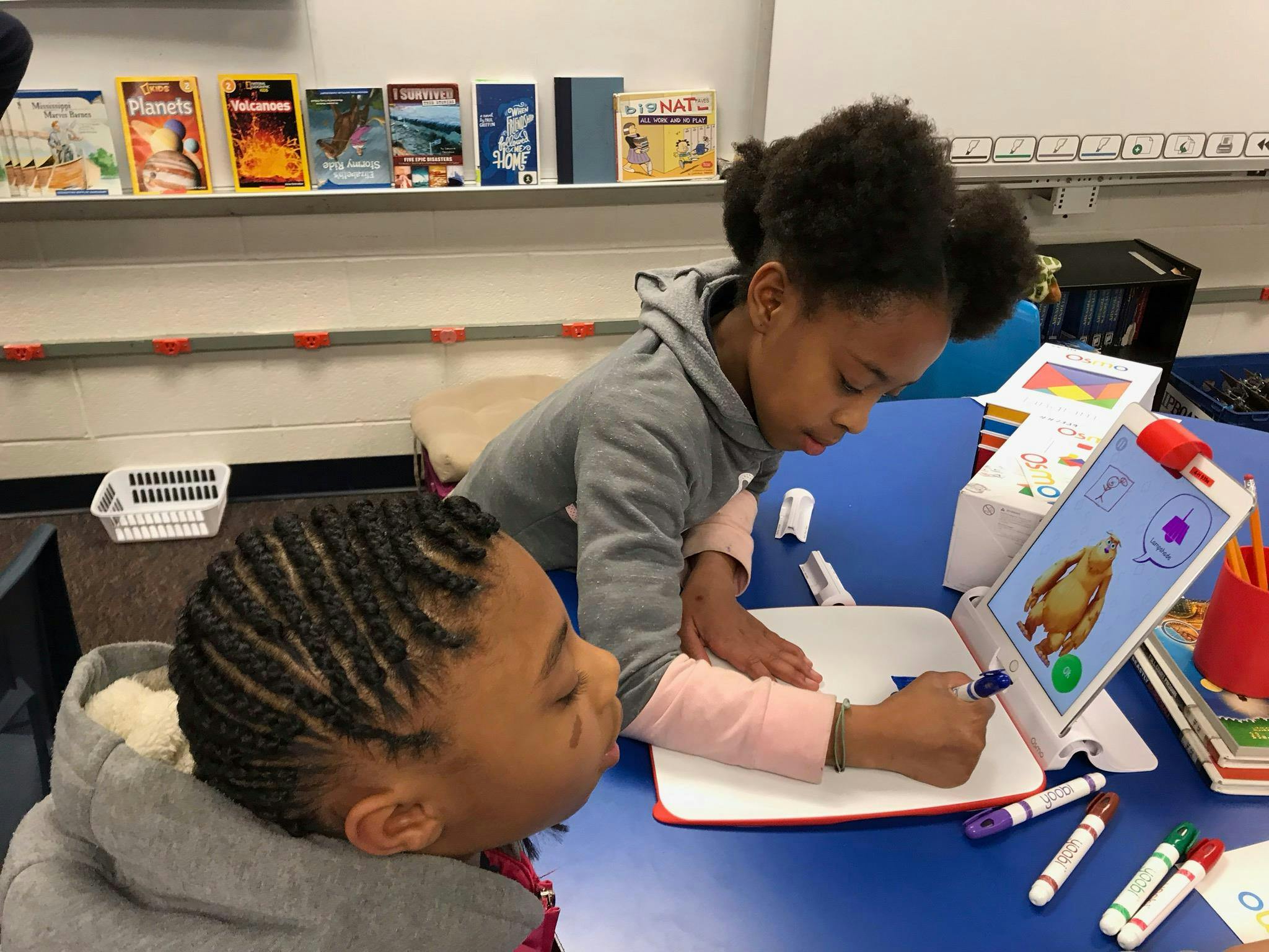 Two children drawing on paper with a tablet in a classroom