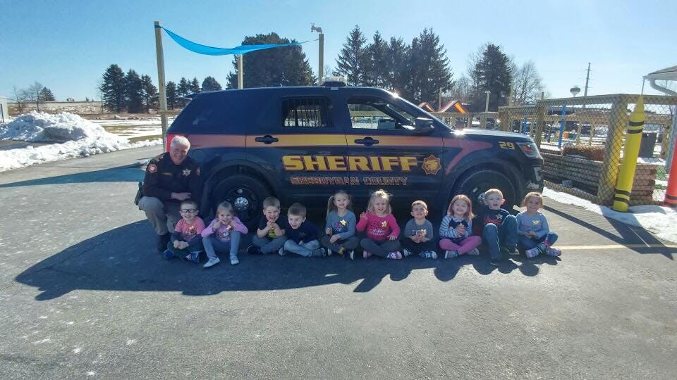 Children sitting in front of a sheriff's vehicle with an adult kneeling beside them