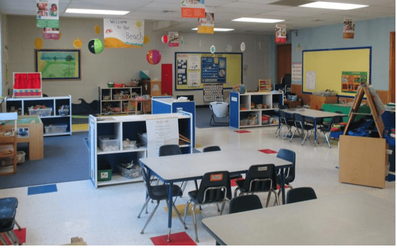Empty classroom with small tables and chairs, shelves with toys