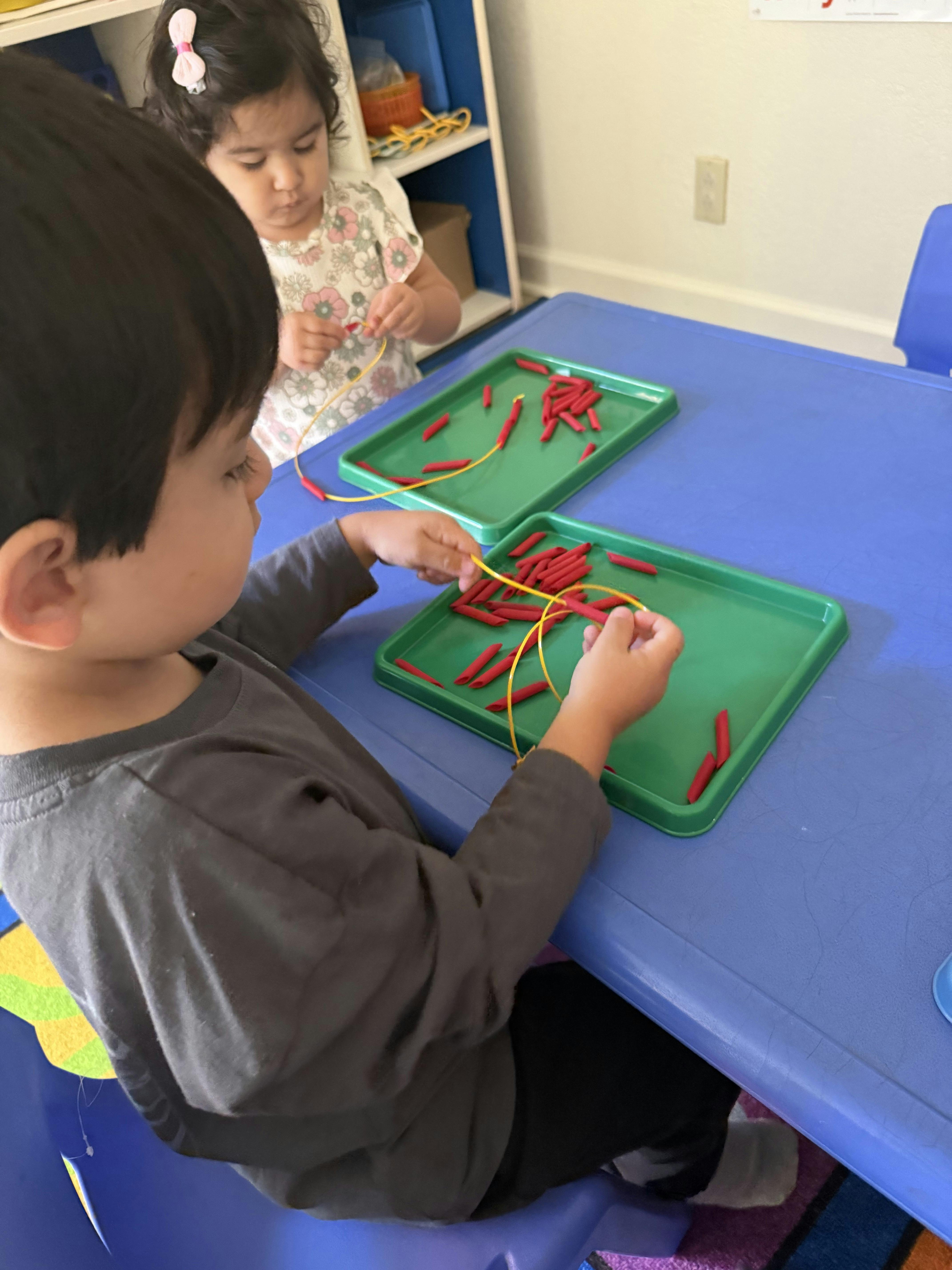 Two children threading pasta pieces onto a string at a table
