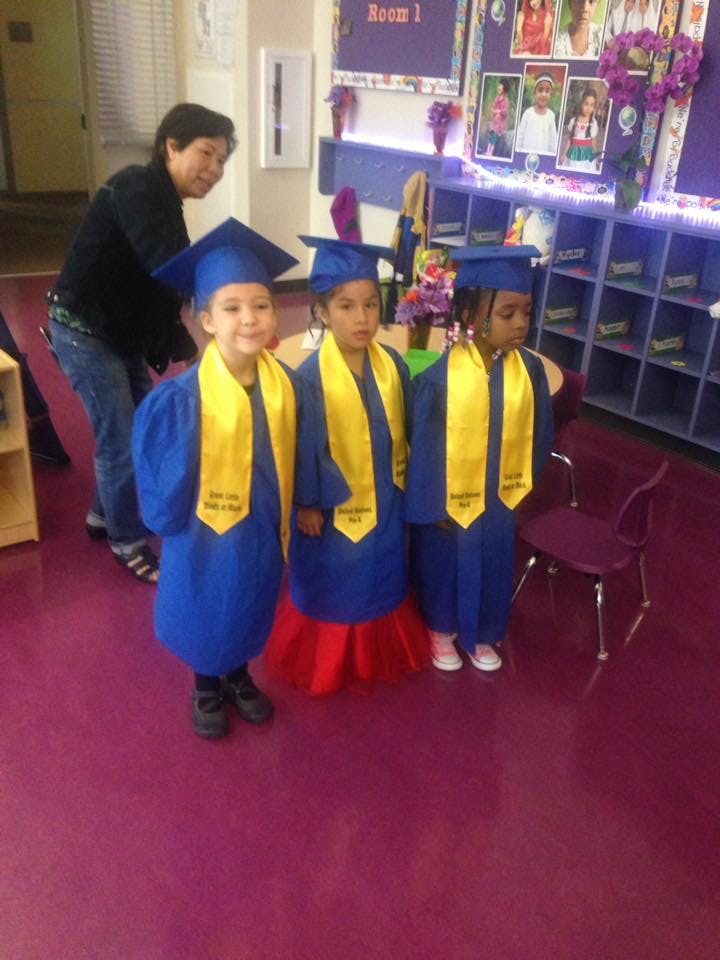 Three children in blue graduation gowns and yellow sashes, with an adult behind them
