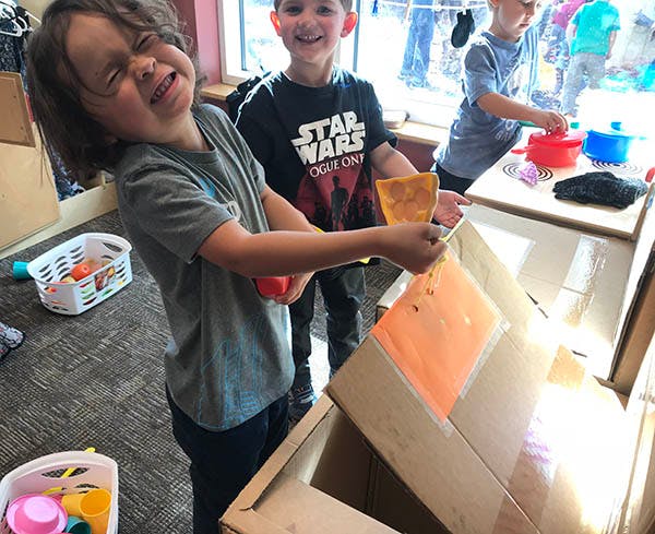 Child playing with a cardboard box and another child smiling in the background