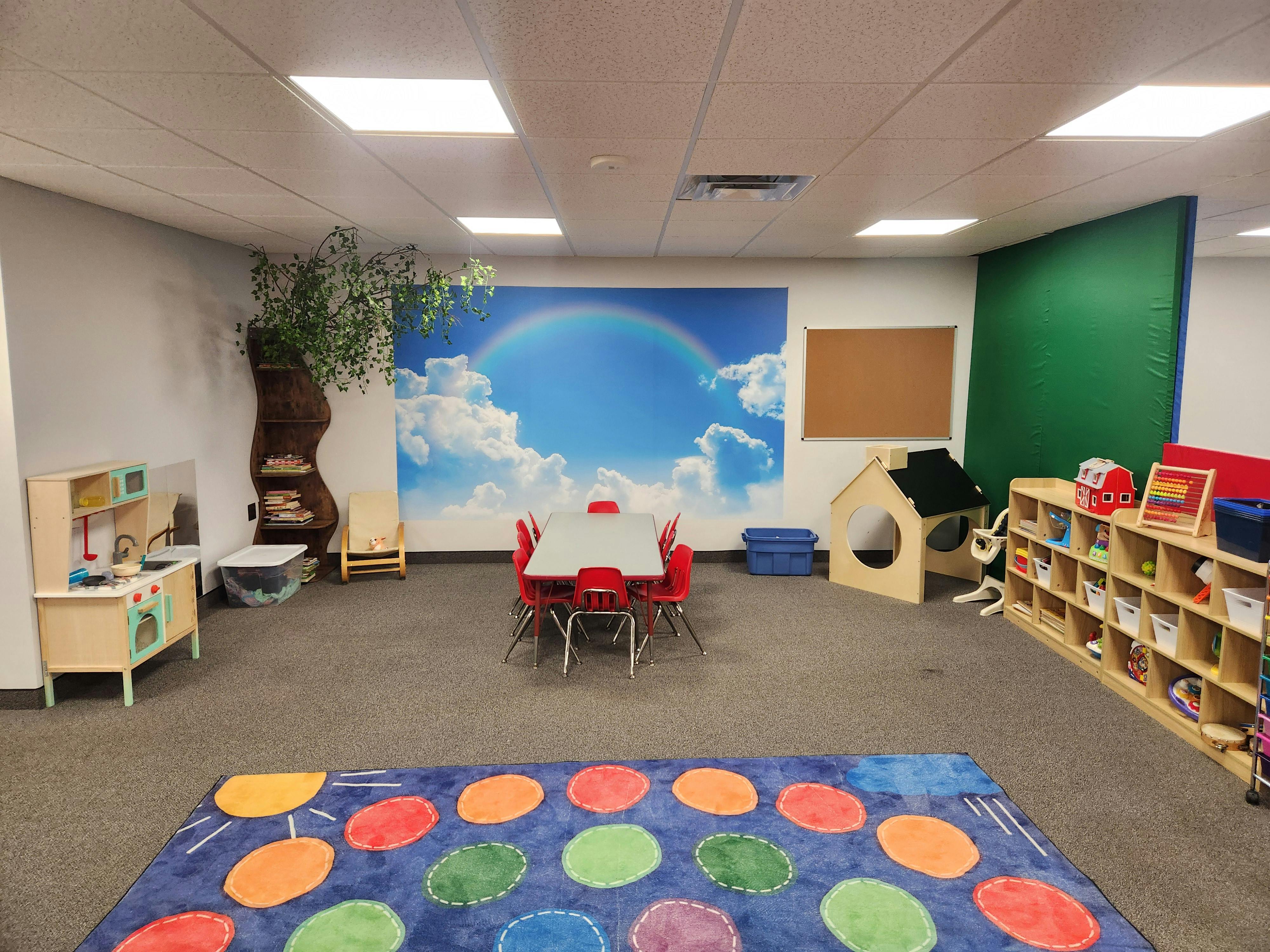 Children's classroom with small table and chairs, play kitchen, and rainbow mural