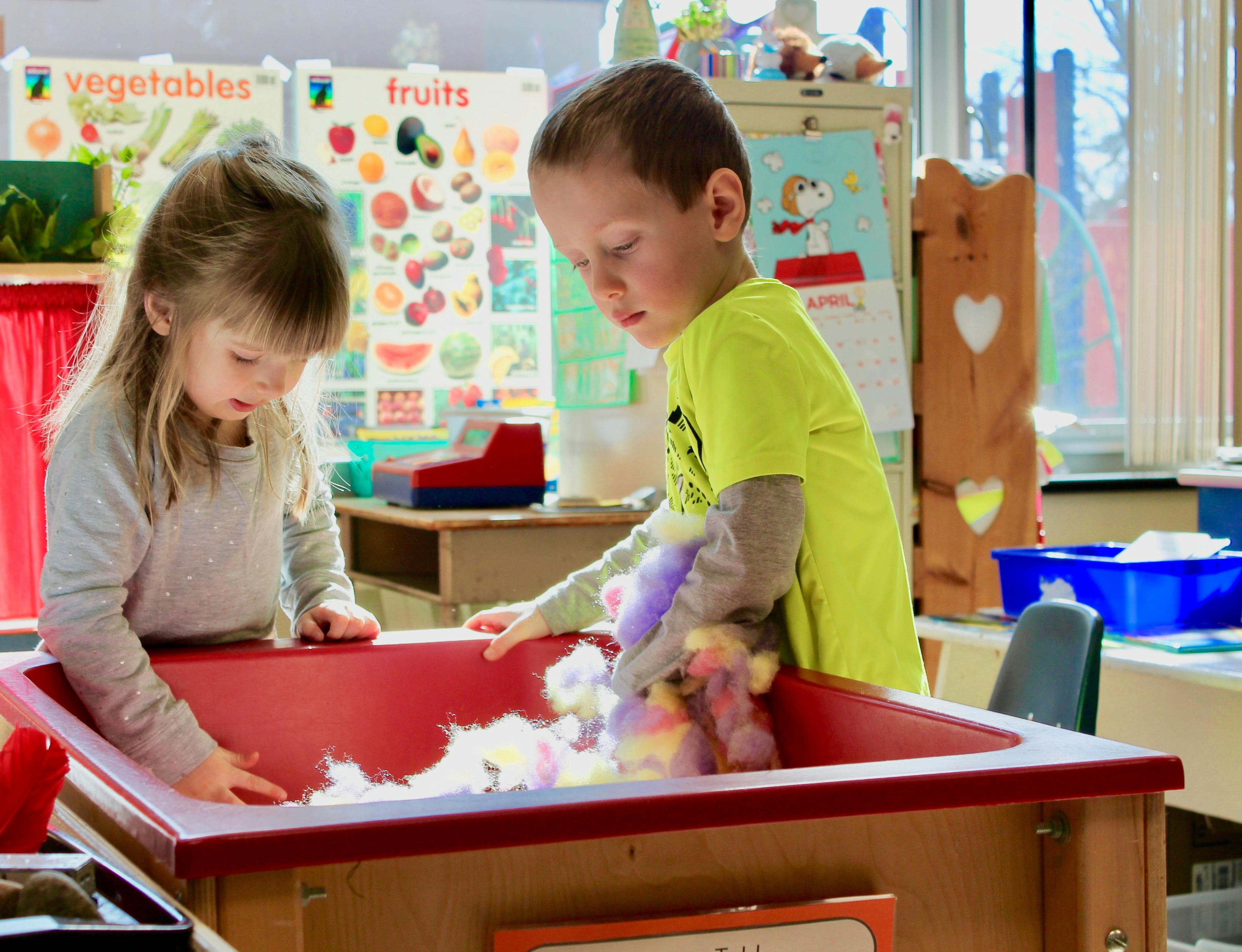 Two children playing with white material in a red bin in a classroom