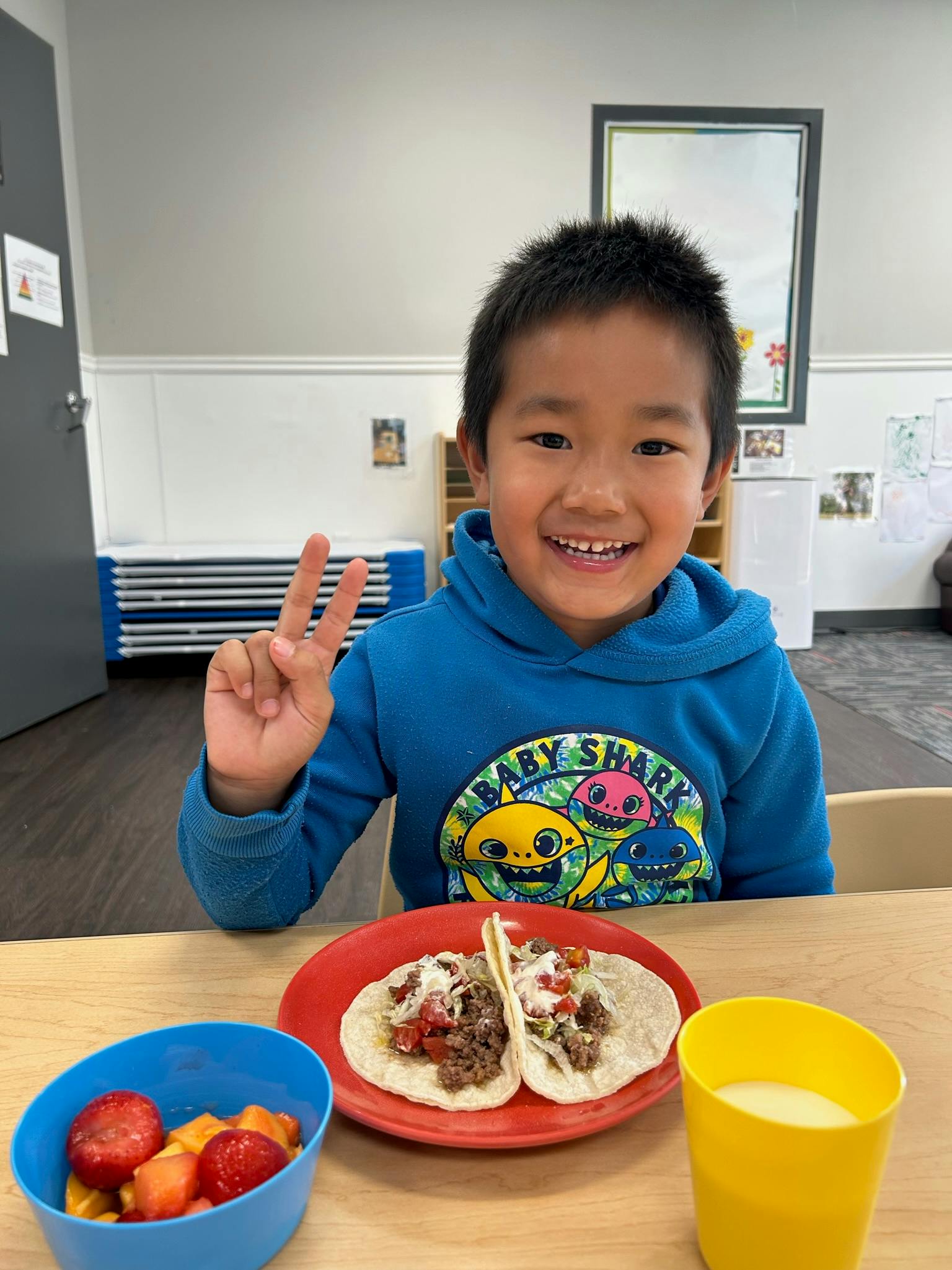 Child making a peace sign at a table with tacos and fruit