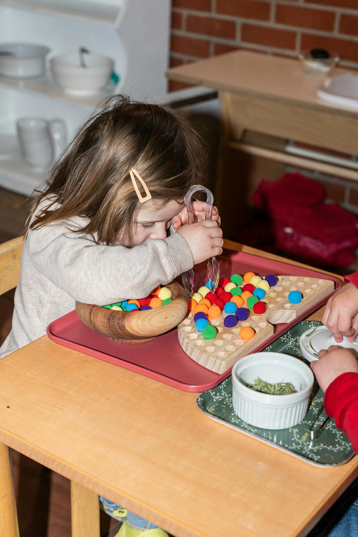 Child using tongs to move colorful pom-poms from a bowl to a board