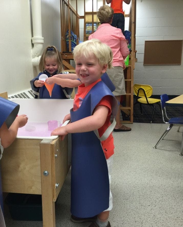 Child in blue smock standing at a water table, another child playing in background