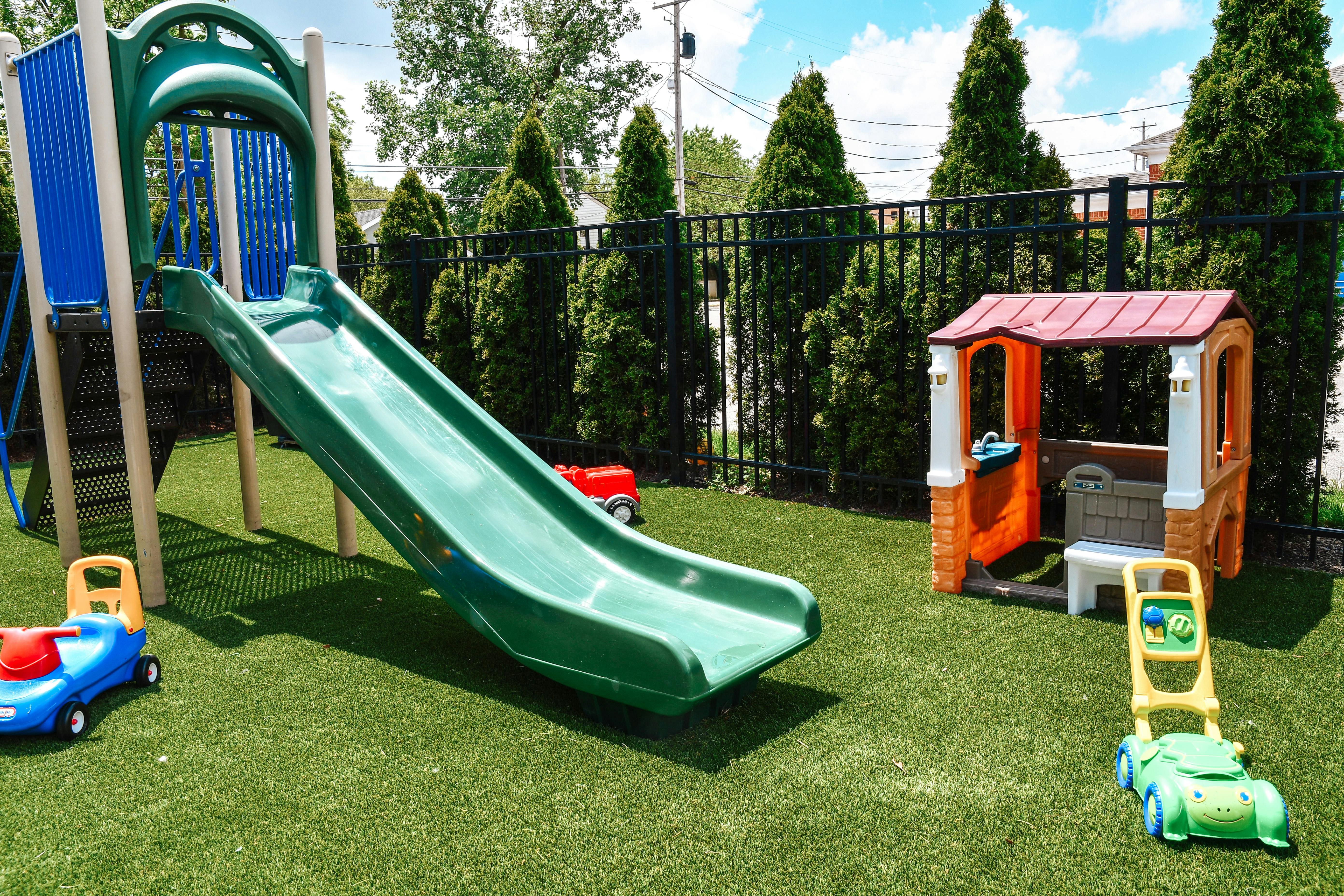 Playground with a green slide, playhouse, and ride-on toys on artificial turf