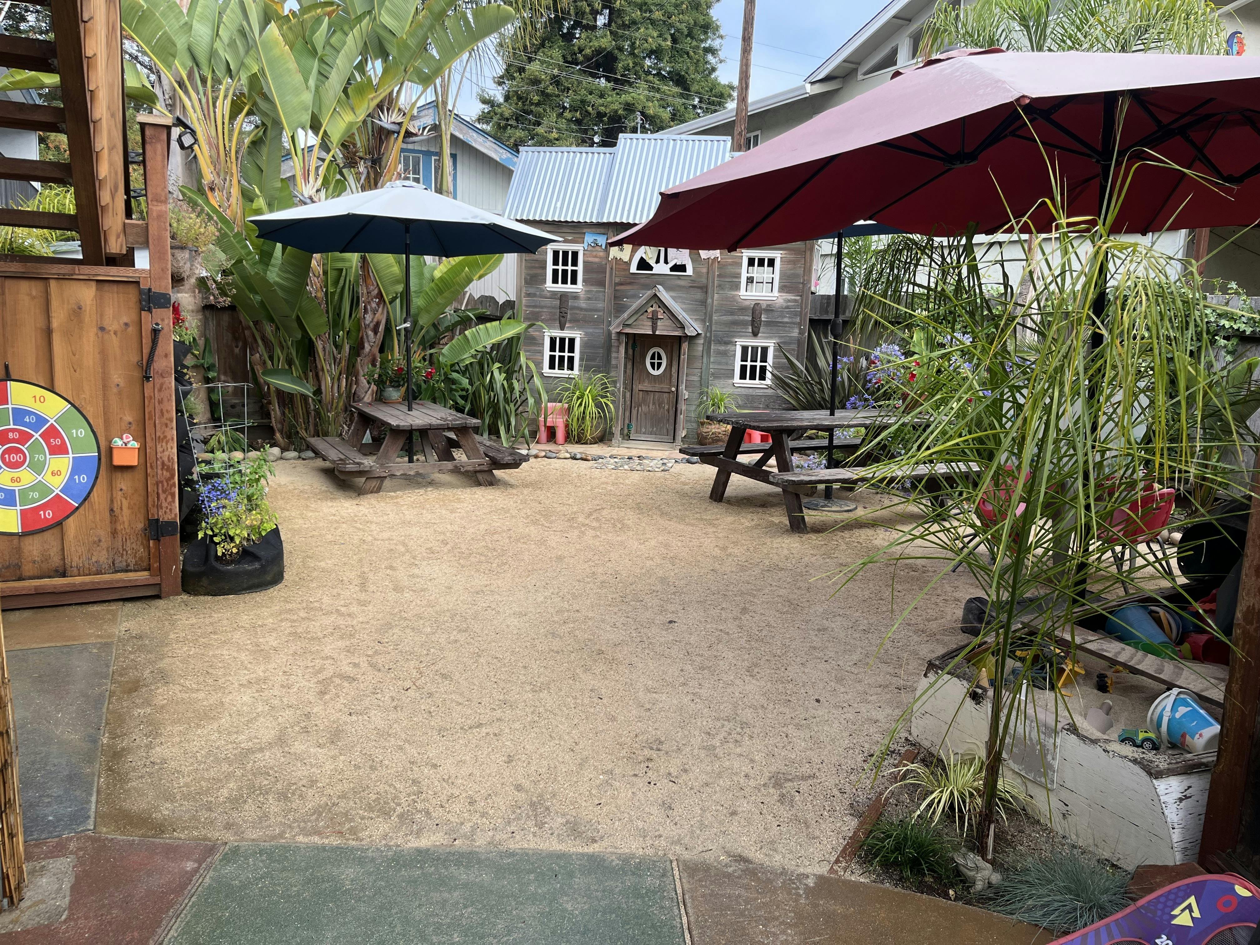 Outdoor play area with a small house, picnic tables, and umbrellas