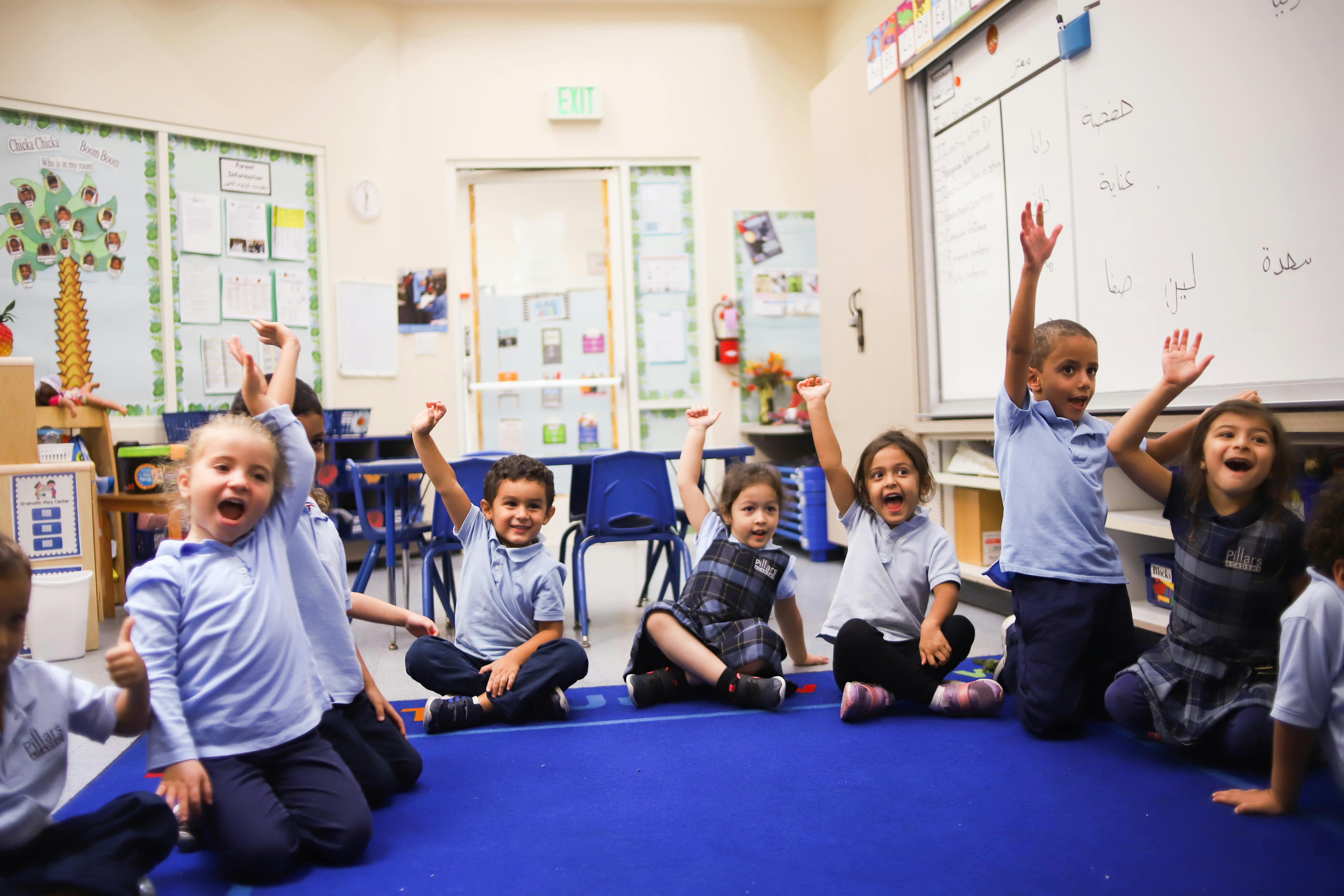 Children sitting on a blue mat in a classroom with hands raised