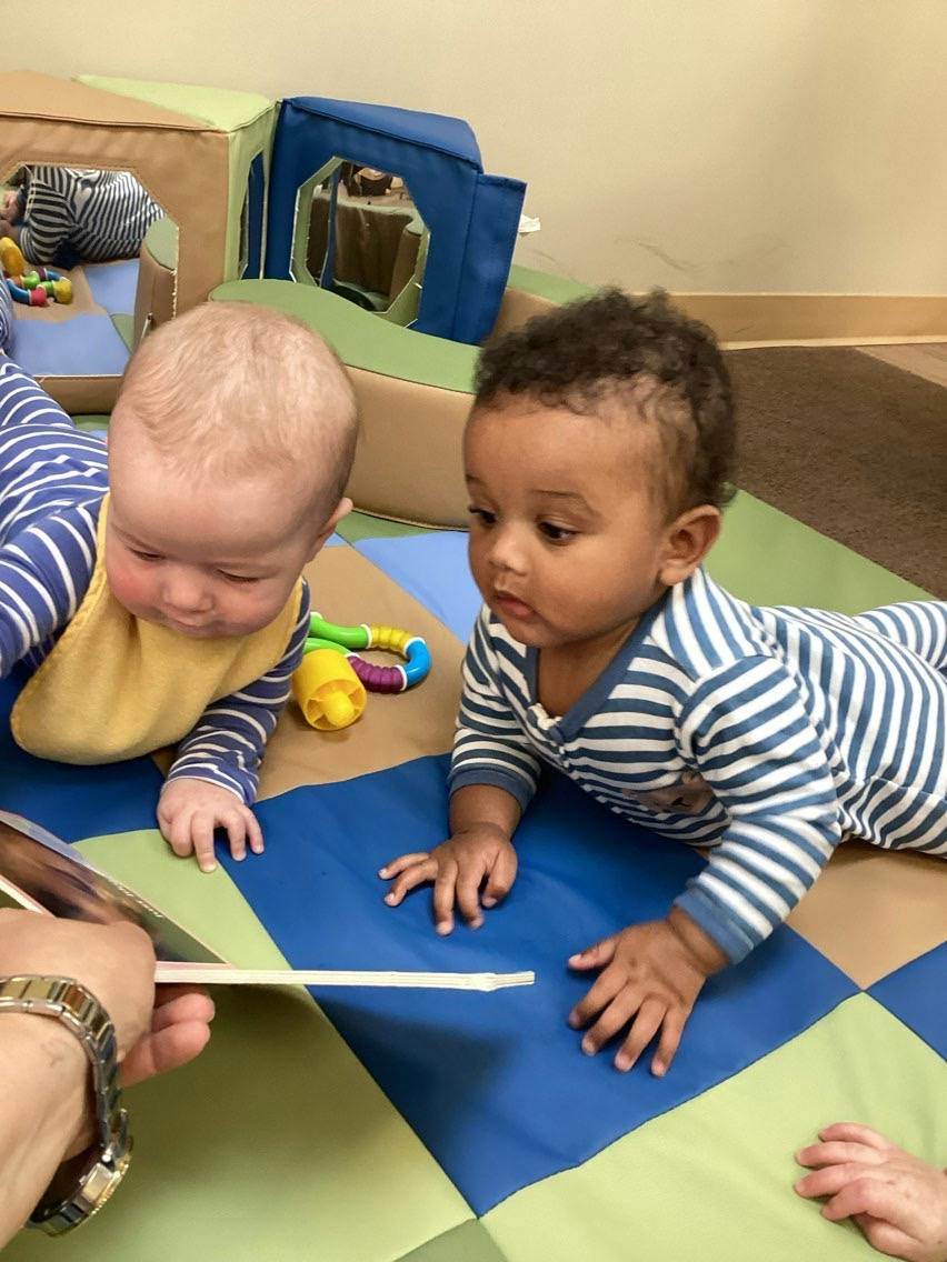 Two children in striped onesies lying on a mat while looking at a book