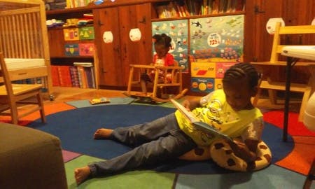 Child reading a book on the floor, another child in a rocking chair
