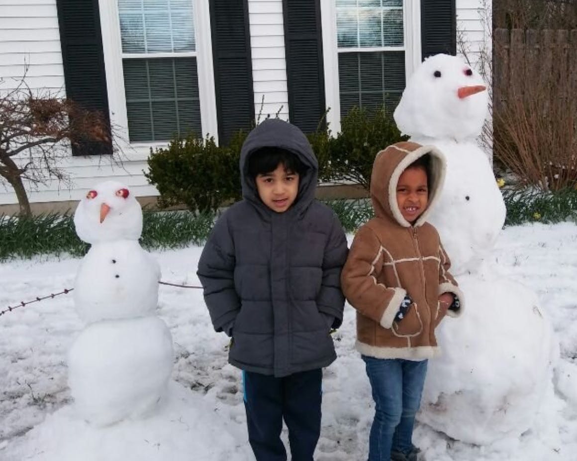 Two children standing in snow next to two snowmen