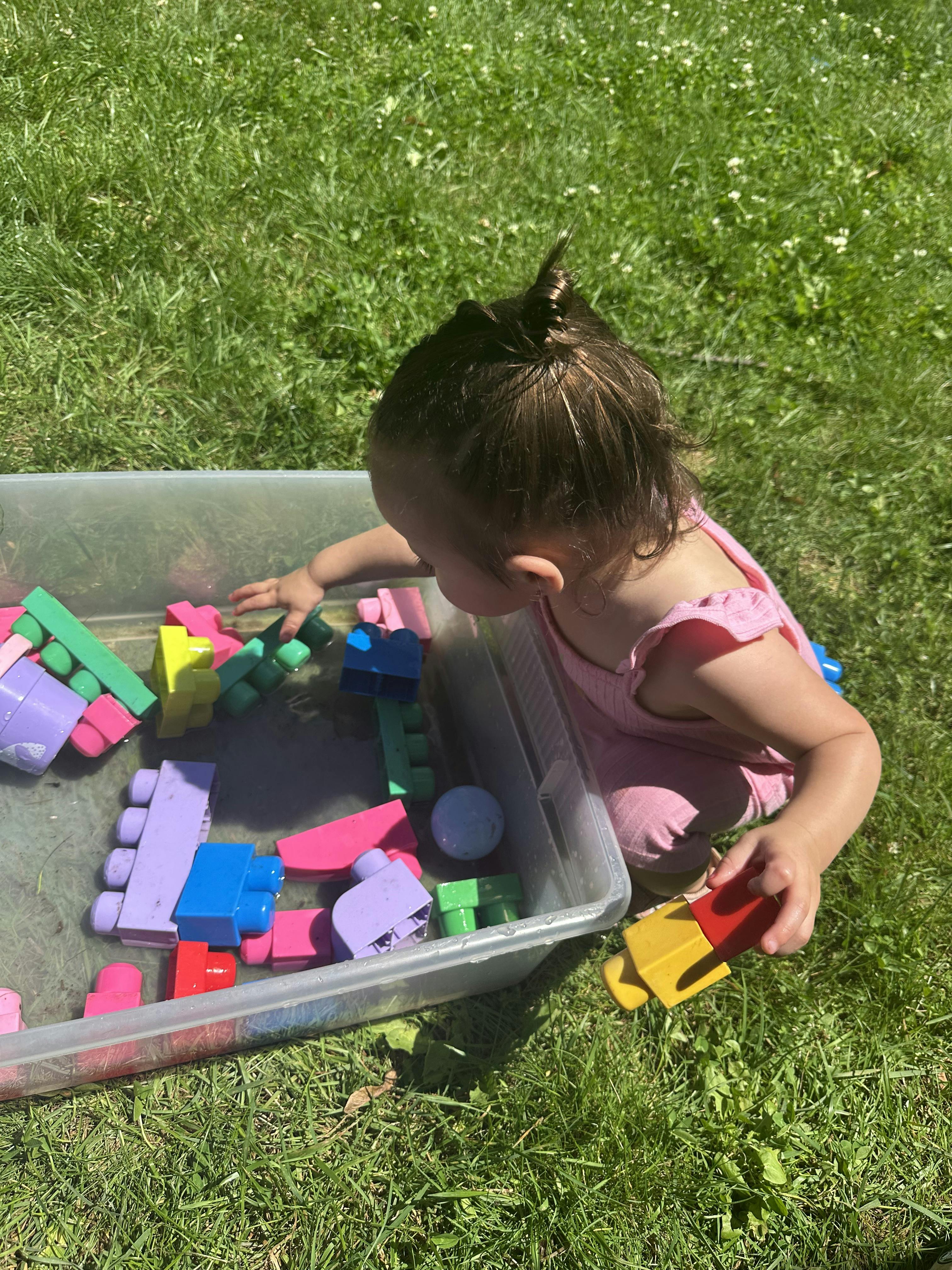 Child playing with colorful blocks in a clear bin on grass