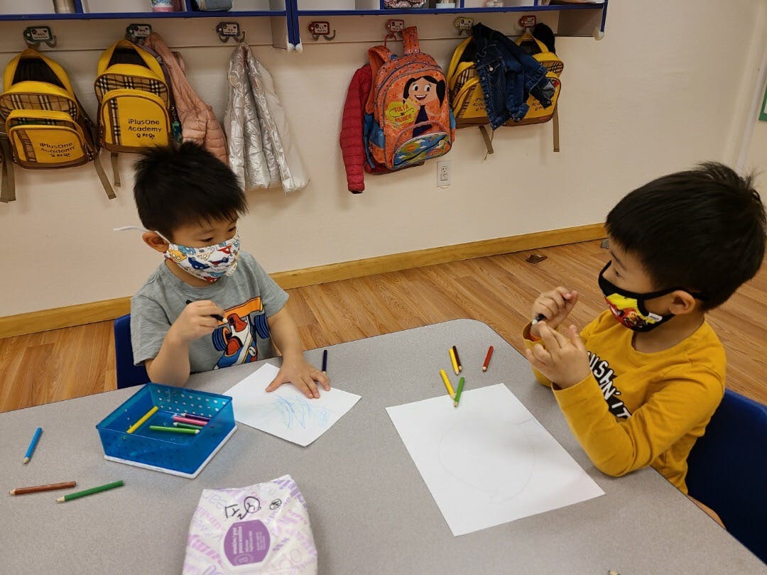 Two children wearing masks drawing at a table