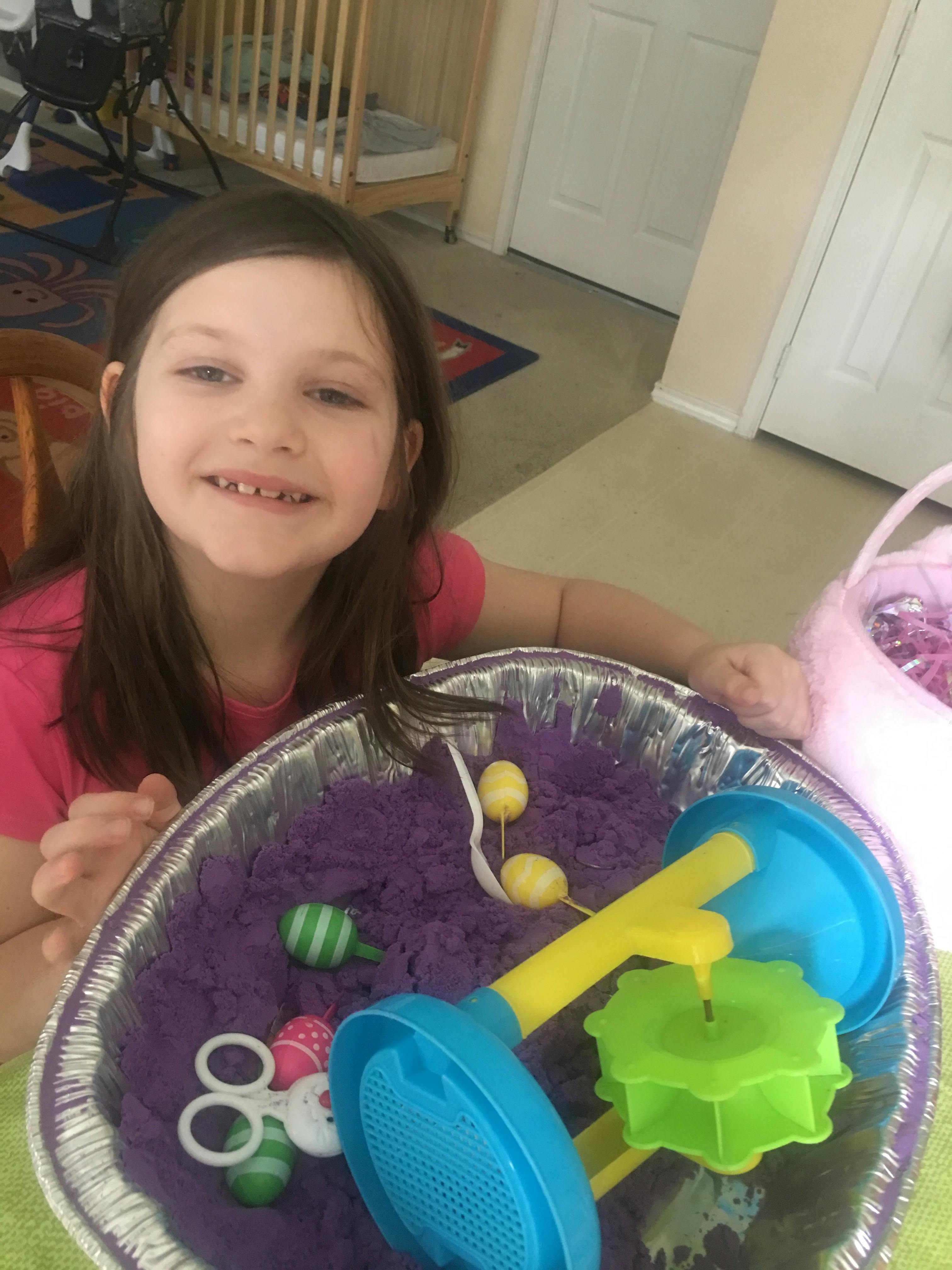 Child smiling next to a tray of purple kinetic sand and toys