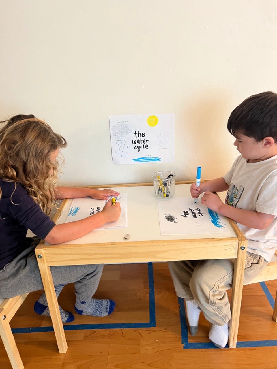 Two children drawing at a table with a sign that says "the water cycle"