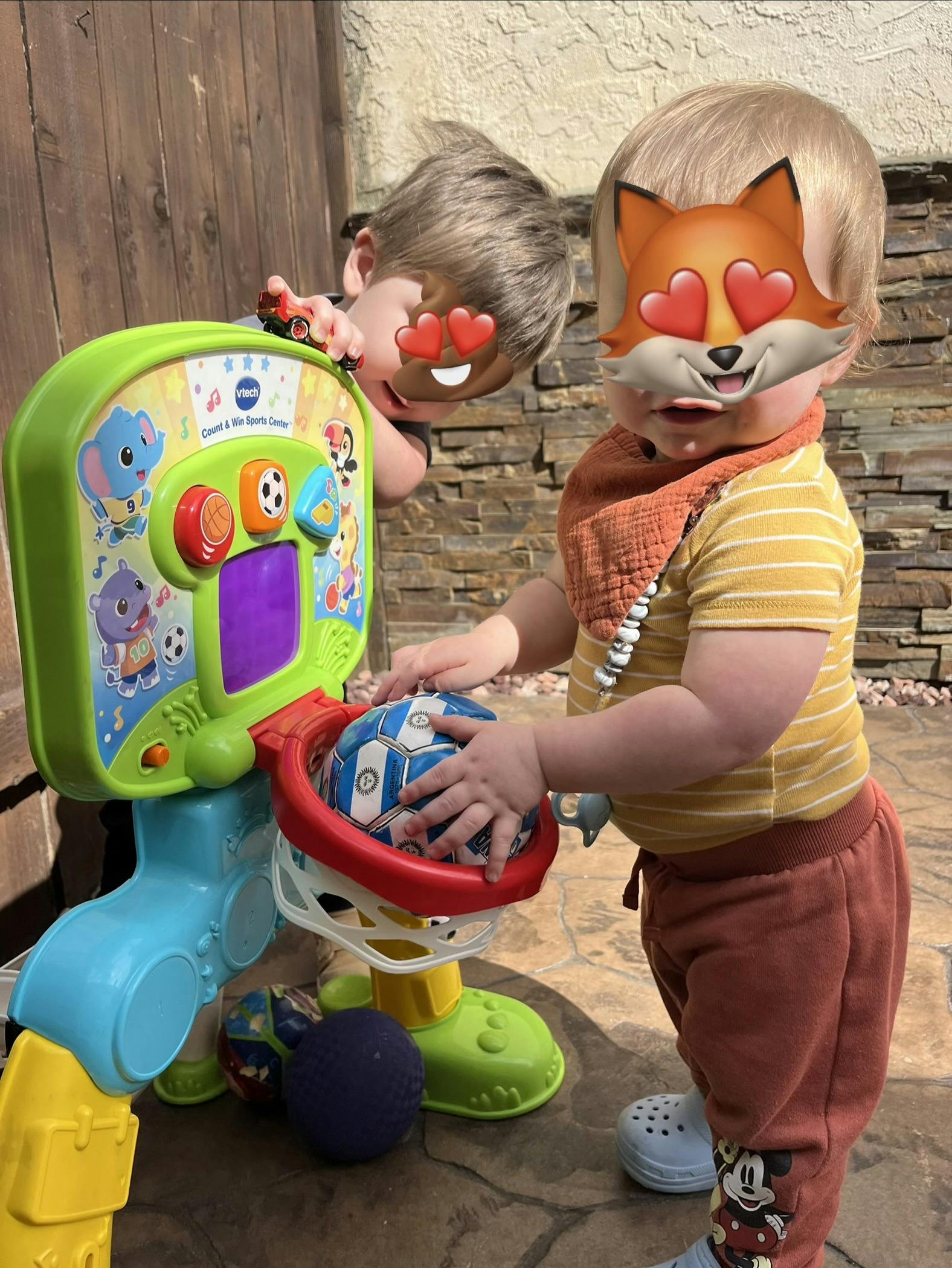 Child placing a soccer ball into a toy basketball hoop, another child playing nearby