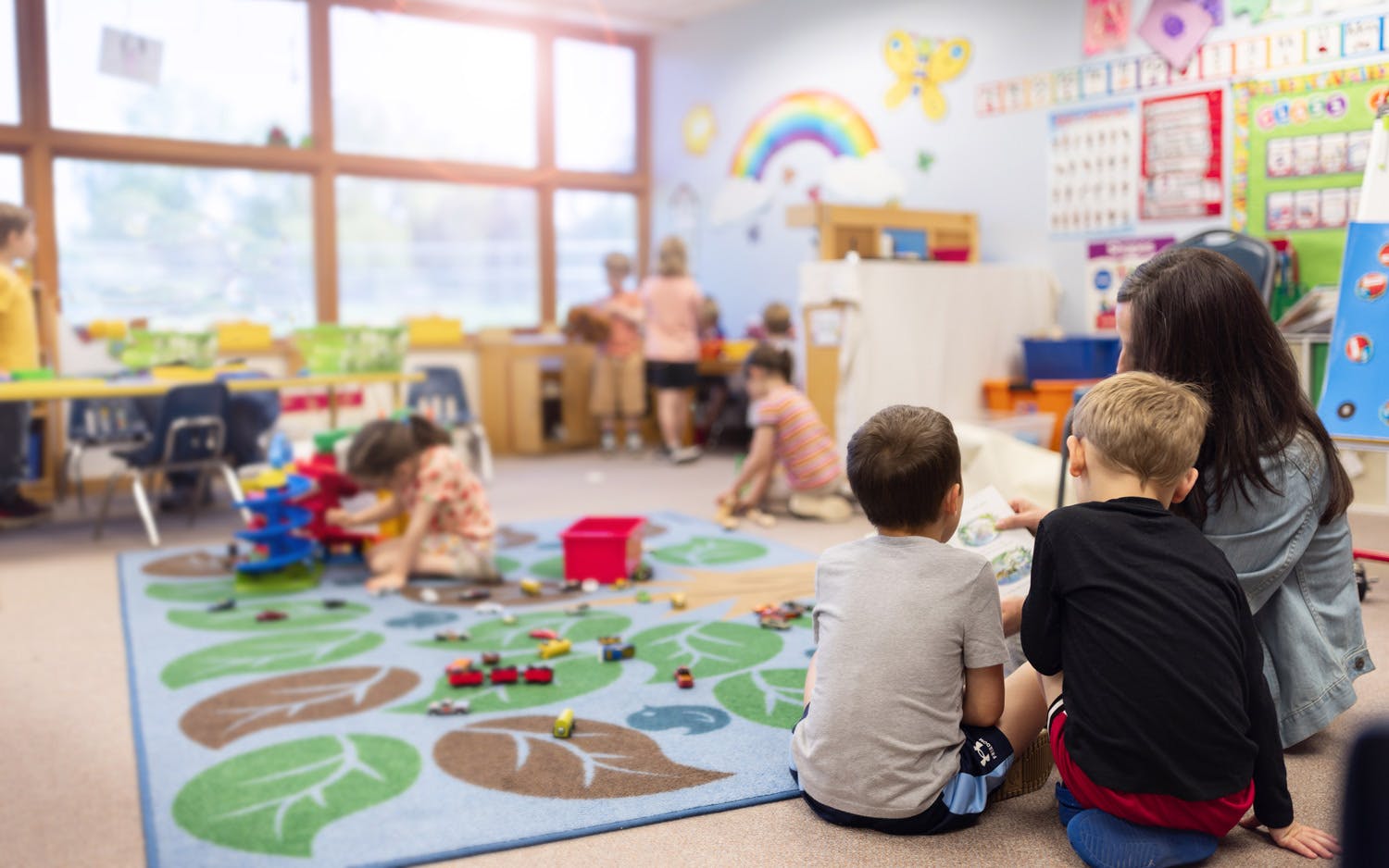 Adult reading to two children in a classroom with other children playing