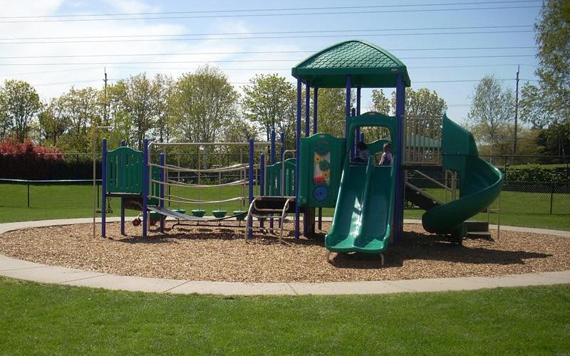 A green and blue playground structure with slides and climbing nets on wood chips, two children playing