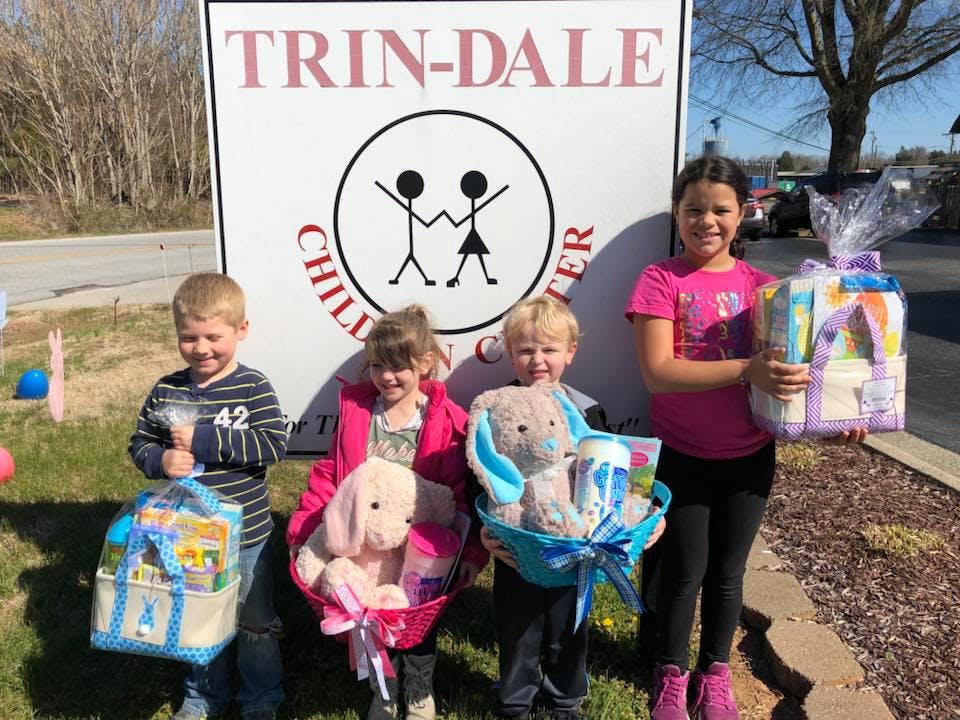 Four children holding baskets with stuffed animals and gifts in front of a sign