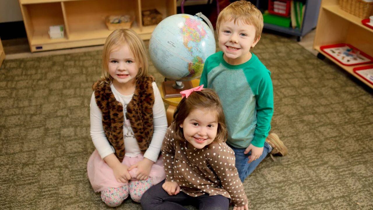 Three children kneeling on a carpeted floor with a globe in a classroom