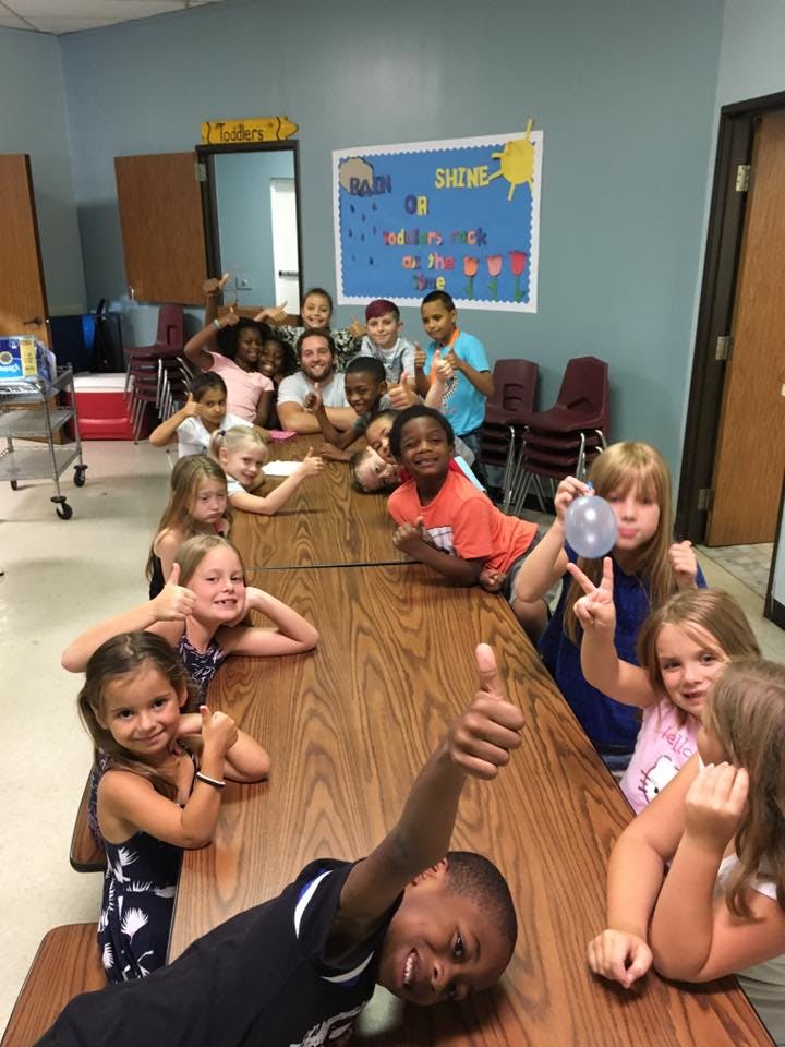 Group of children and one adult sitting at a long table, giving thumbs up