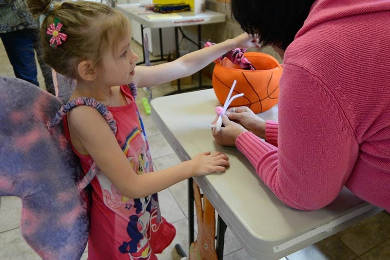 Child in fairy wings reaching for an orange basket while a person works on a craft
