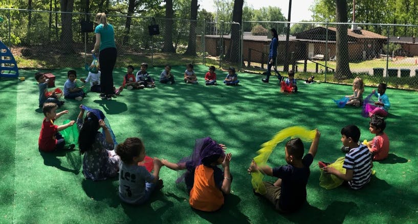Children sitting in a circle on green turf with an adult standing