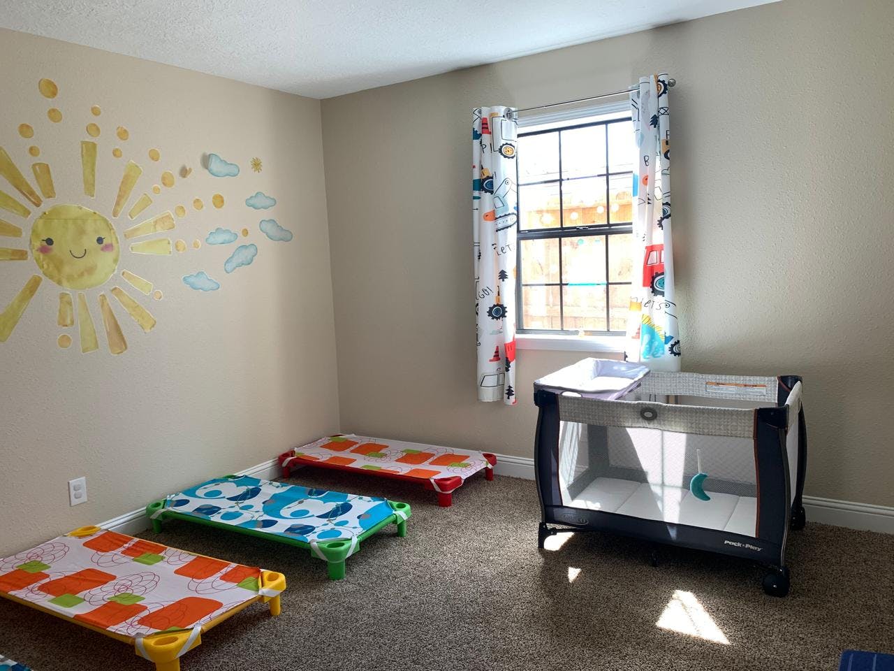 Childcare room with three cots, a playpen, and a sun mural on the wall