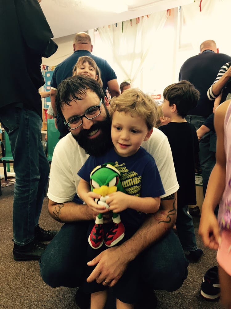 Person with beard and glasses holding a child with a stuffed toy