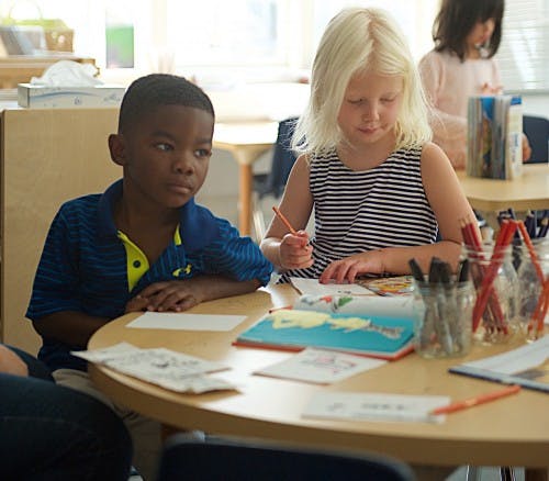Two children at a table, one drawing and one looking away