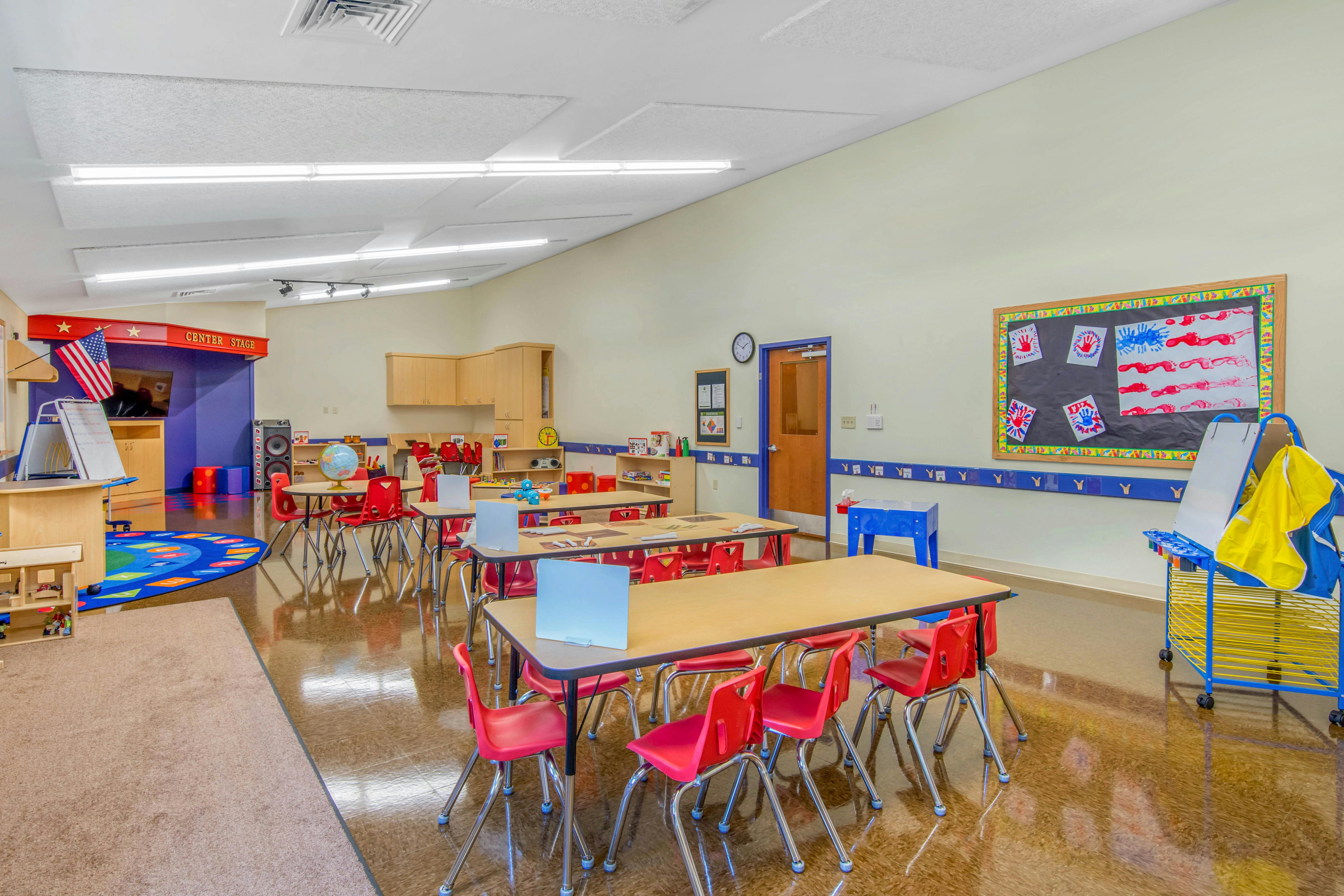 Classroom with small tables and red chairs, a stage, and activity rug