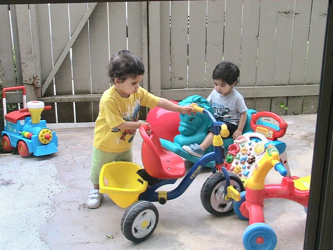 Two children with tricycles and other toys in a yard