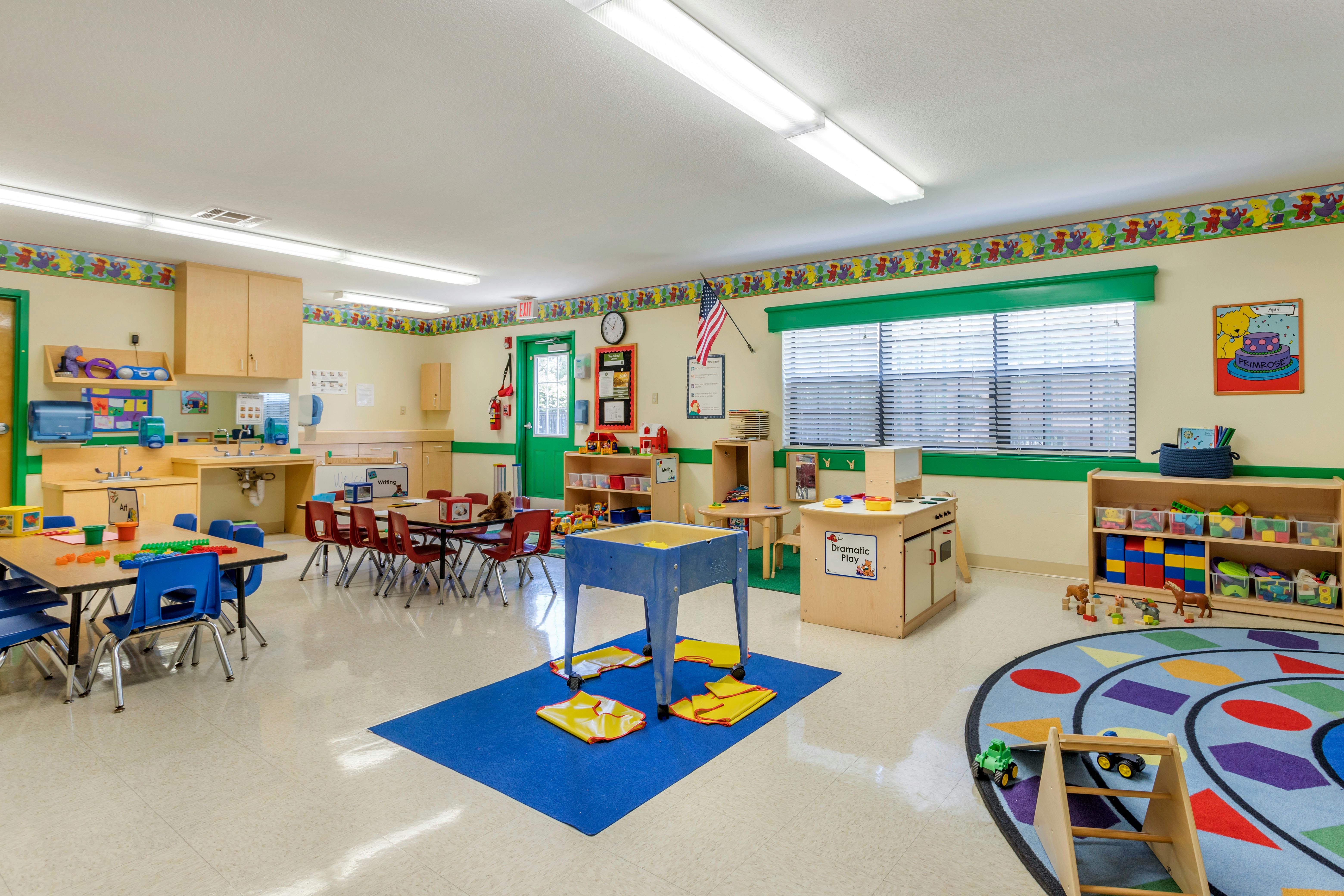 A colorful empty classroom with small tables and chairs, a play kitchen, and storage shelves