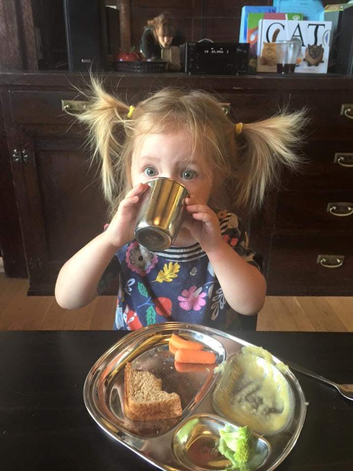 Child drinking from a metal cup at a table with a divided plate of food