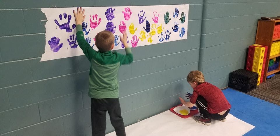 Child making handprints on paper, another child painting on floor with paint