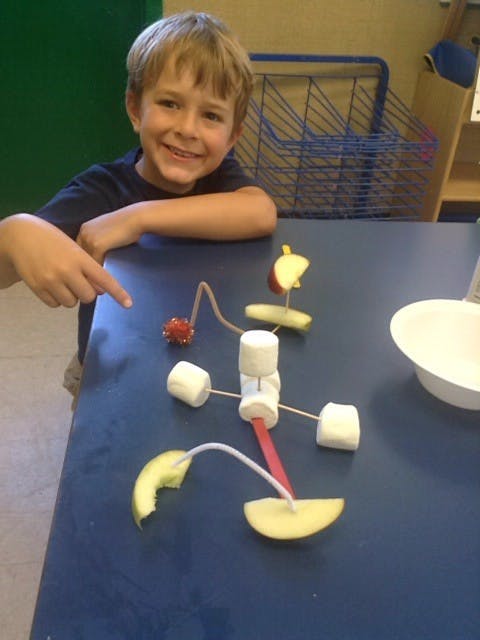 Child pointing at a food sculpture made of marshmallows, apple slices, and sticks