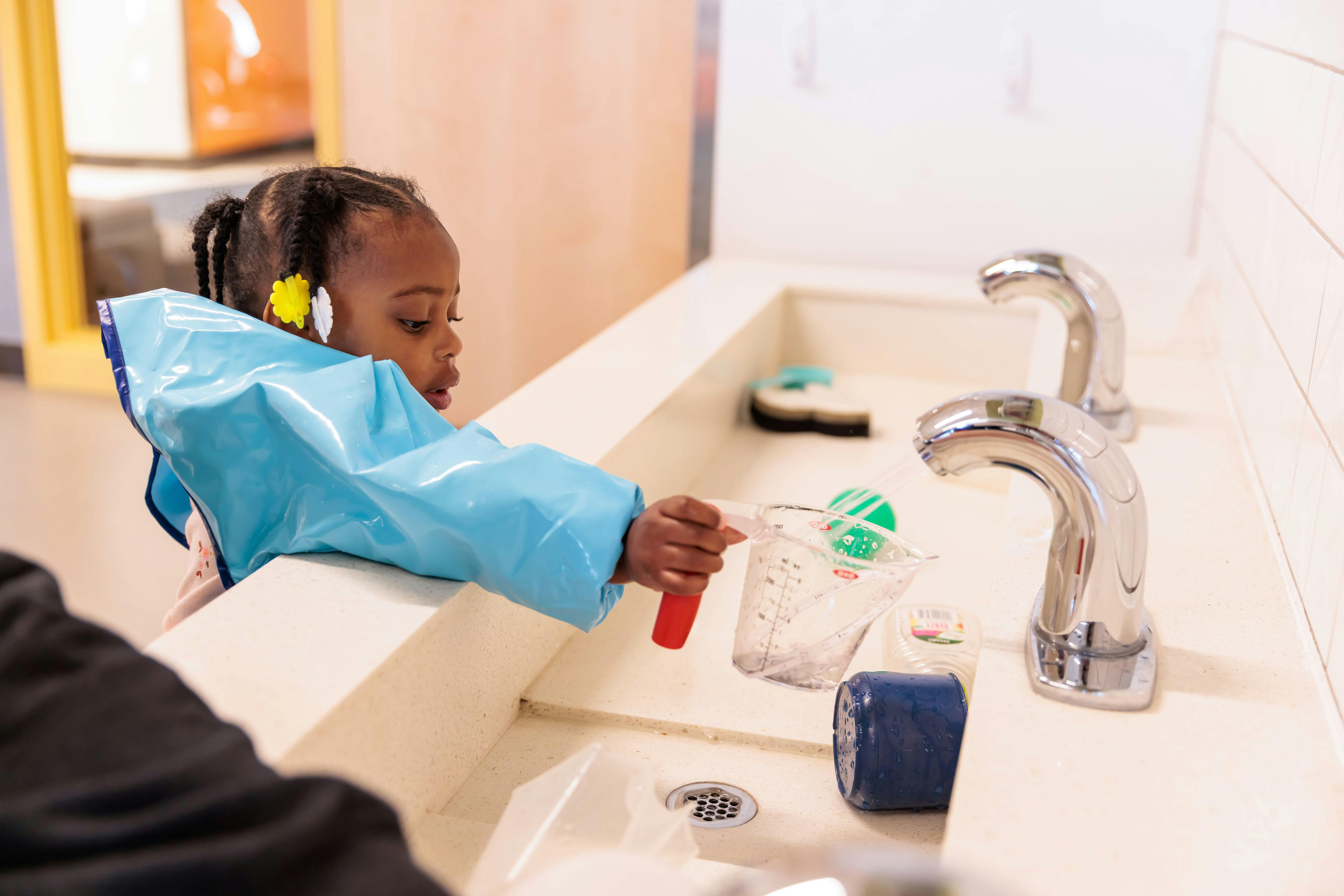 Child in a blue smock playing with a cup and a red tool at a sink