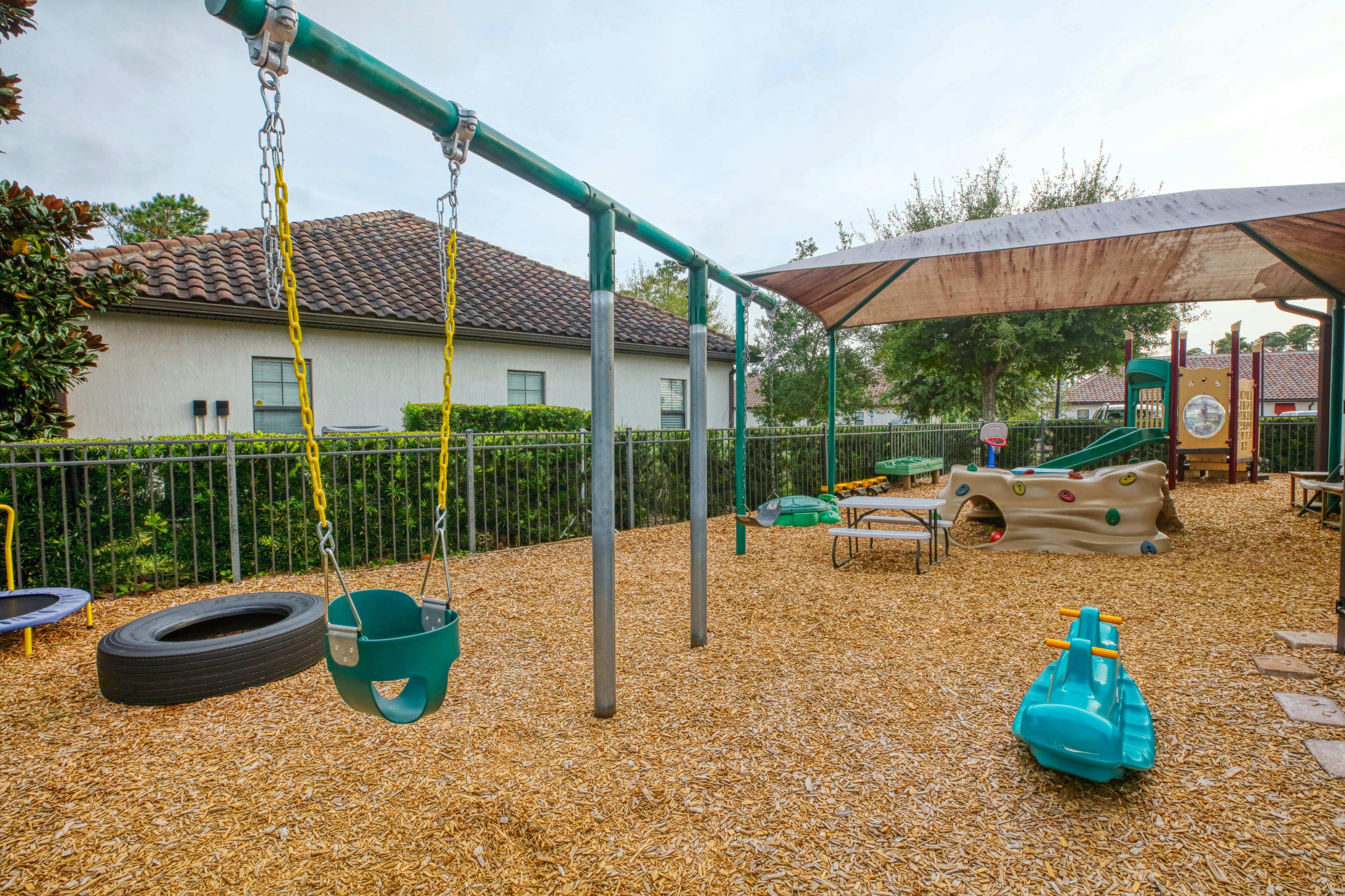 An outdoor playground with swings, a slide, climbing wall, picnic table, and wood chips