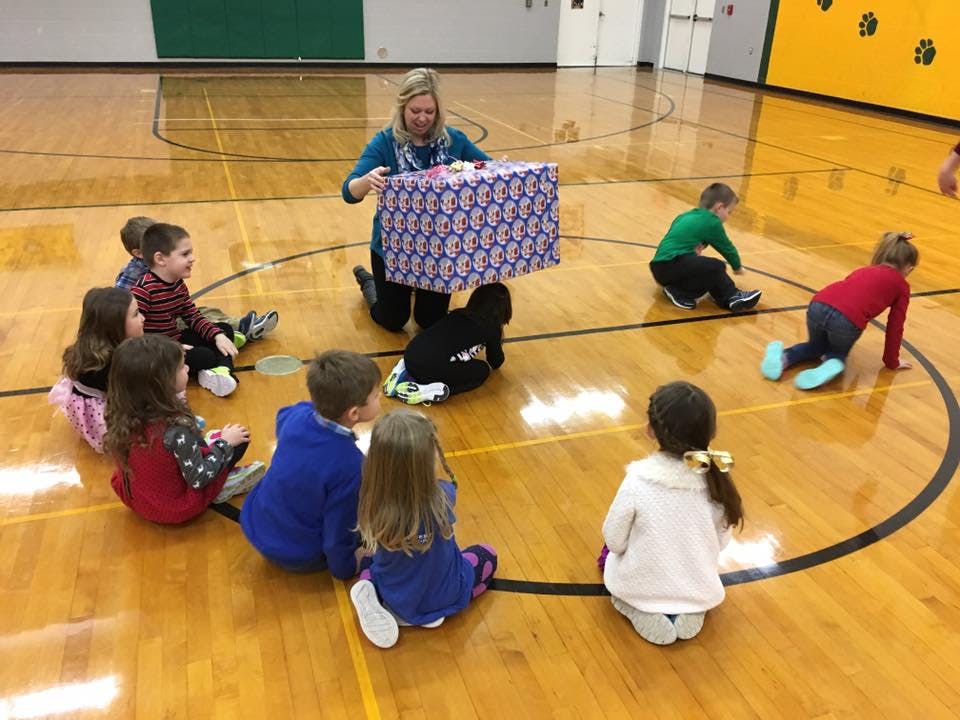 Adult holding a large wrapped gift for children sitting in a circle on a gym floor