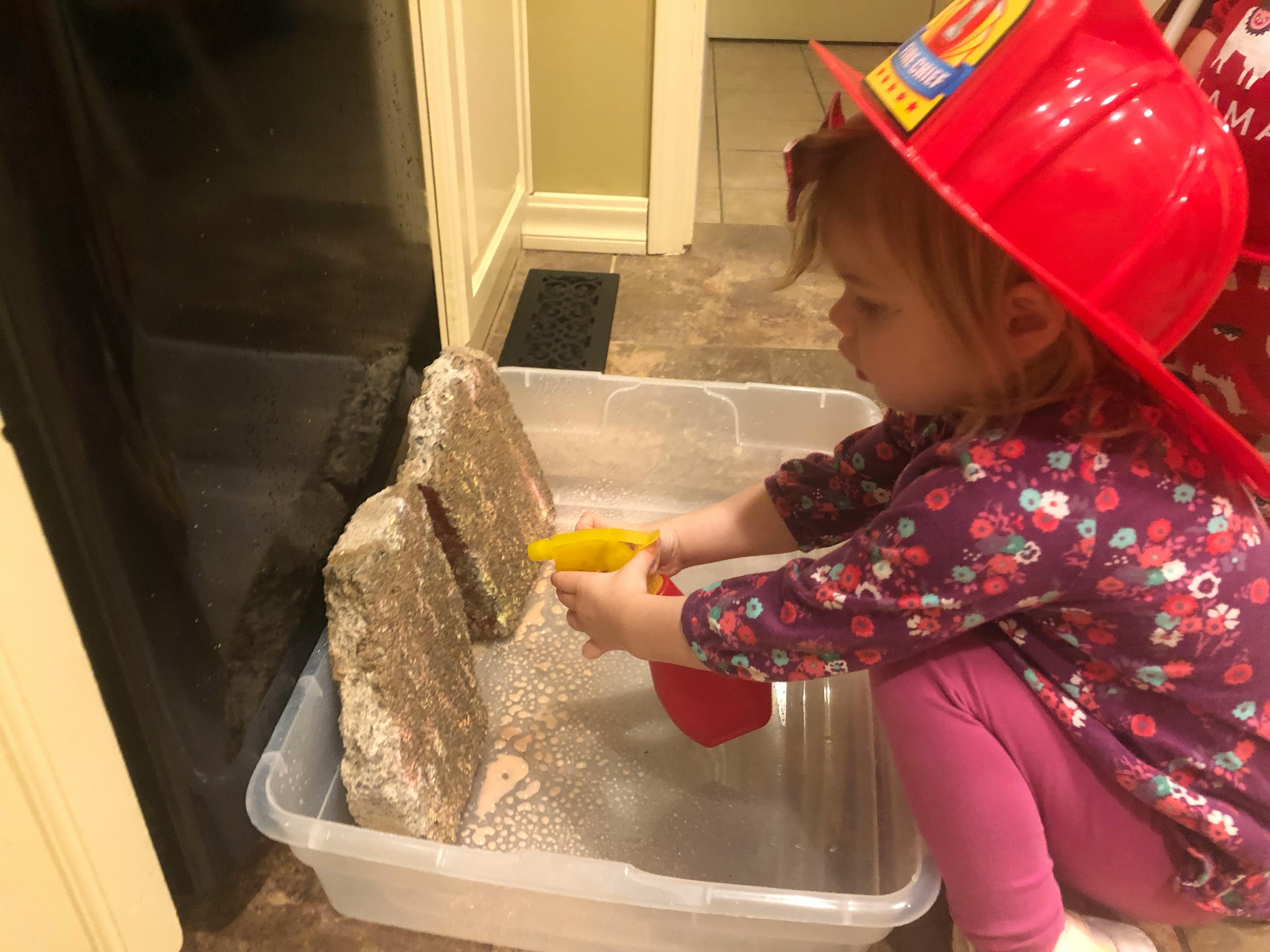 Child in a firefighter hat spraying water on rocks in a bin