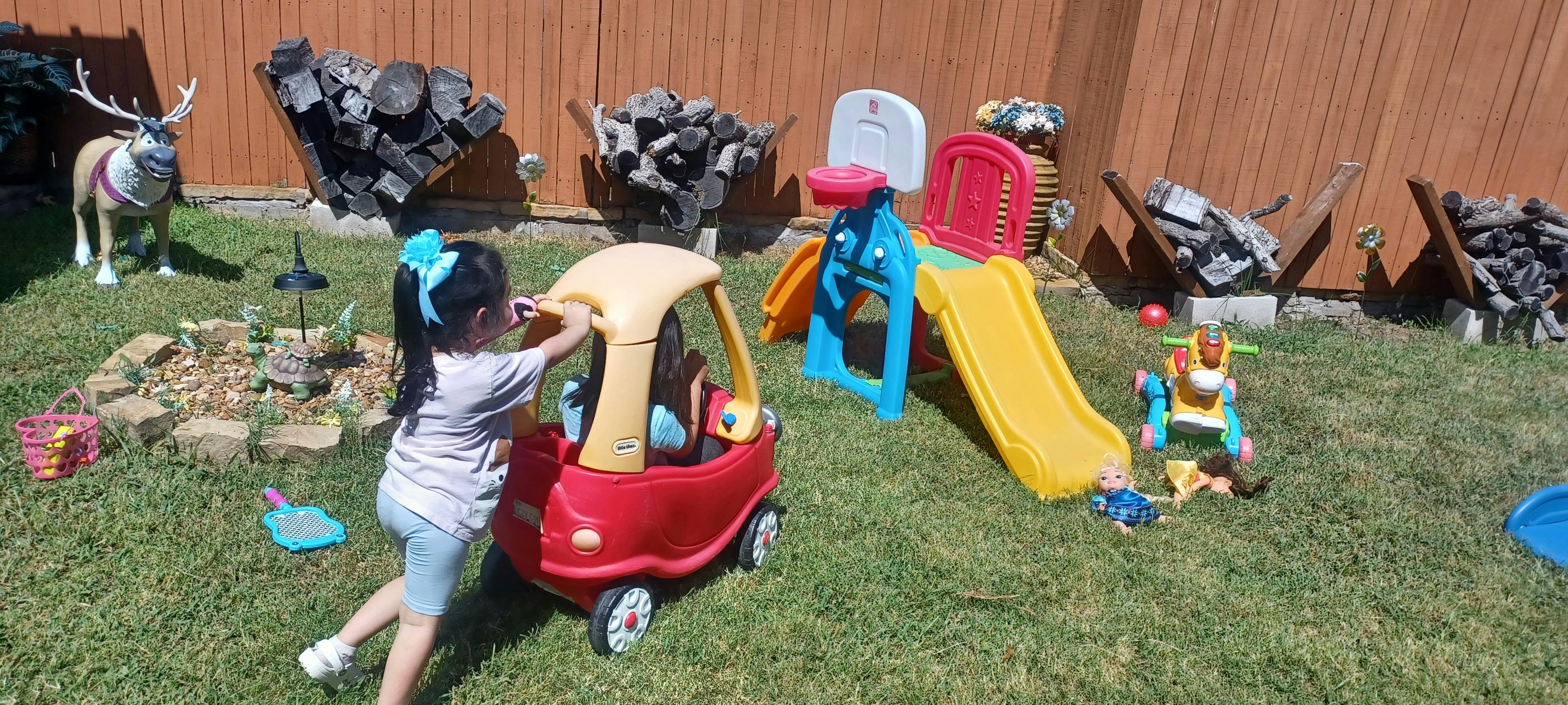 Child pushing a toy car with another child inside on a backyard lawn