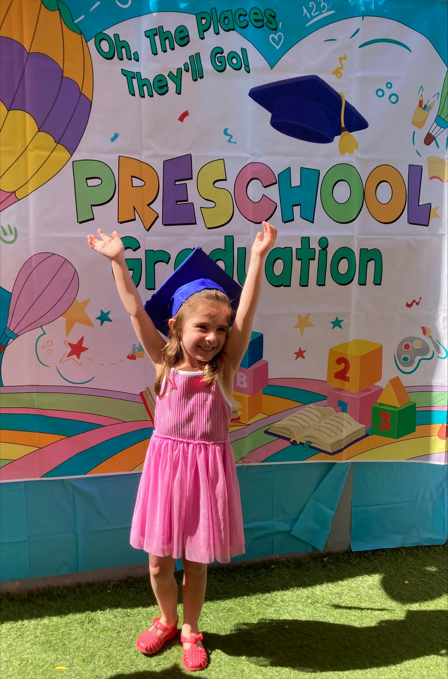 Child in a pink dress and graduation cap with arms raised in front of a banner