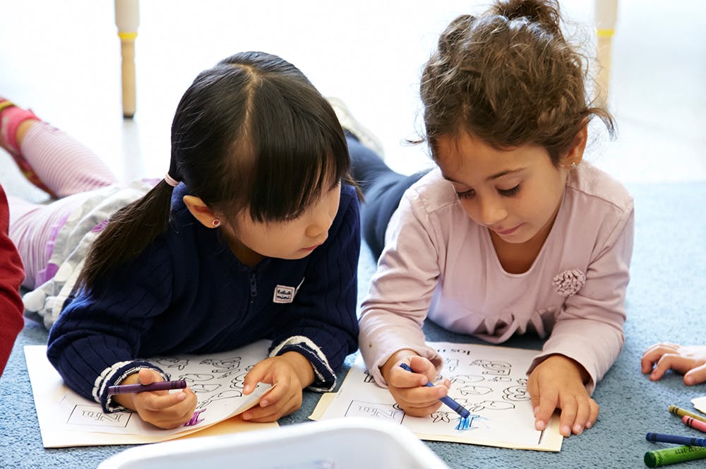 Two children lying on the floor coloring in a classroom