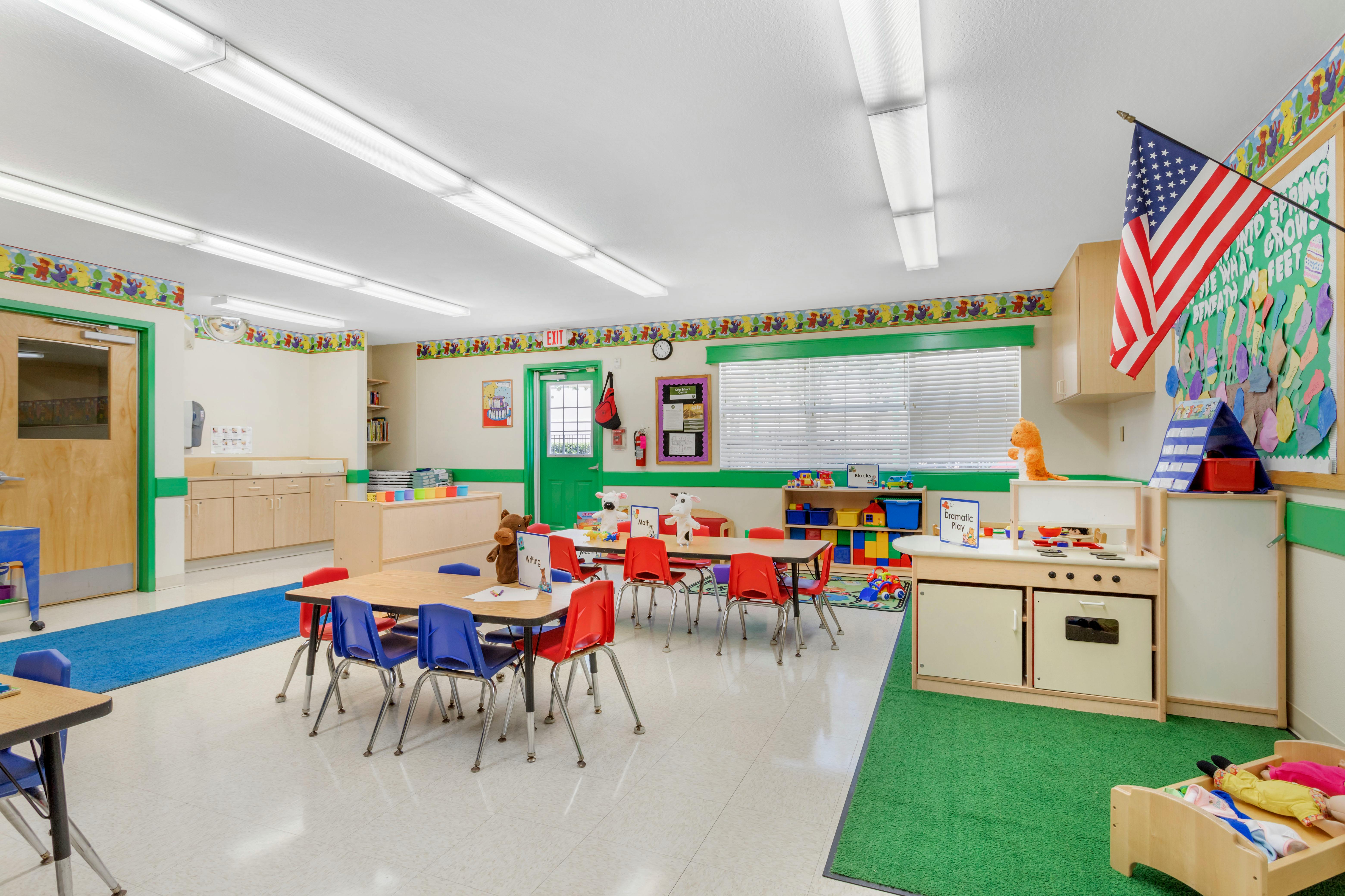 An empty children's classroom with small tables and chairs, toys, and a flag